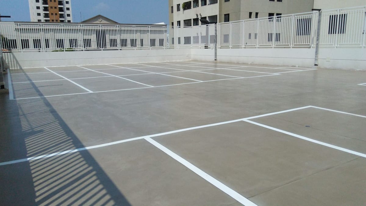 Vista ampla do estacionamento descoberto, com vagas demarcadas e o gradil do edifício ao fundo